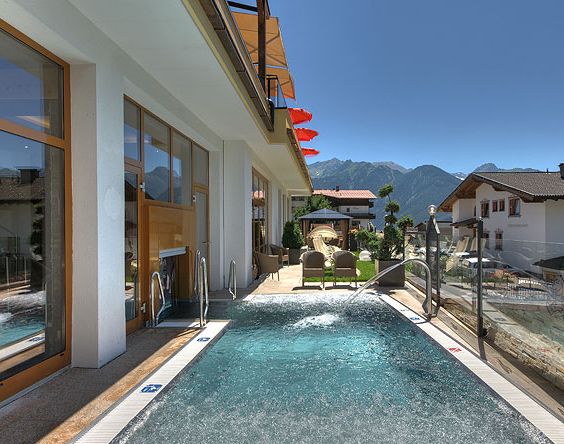 Außenpool neben einem modernen Gebäude mit Liegestühlen und roten Sonnenschirmen vor der Kulisse der Berge und des klaren blauen Himmels.