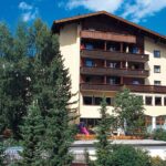 Ein mehrstöckiges Gebäude mit Holzbalkonen, umgeben von Bäumen, vor einem blauen Himmel mit vereinzelten Wolken.