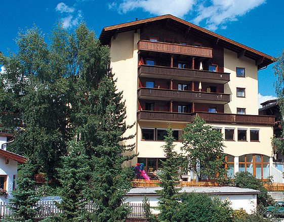 Ein mehrstöckiges Gebäude mit Holzbalkonen, umgeben von Bäumen, vor einem blauen Himmel mit vereinzelten Wolken.