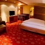 Ein Hotelzimmer mit einem Doppelbett, einem Sofa, einem Fernseher auf einer Holzkommode, einem kleinen Esstisch mit Stühlen und einem gemusterten roten Teppichboden.