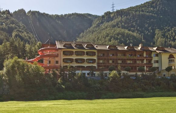 Ein mehrstöckiges Hotel mit Balkonen und einer roten Spiralrutsche vor einem bewaldeten Hügel im Sonnenlicht.
