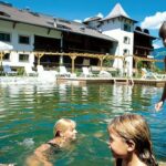 Vier Kinder schwimmen und spielen an einem sonnigen Tag in einem natürlichen Außenpool mit einem Hotelgebäude und Liegestühlen im Hintergrund.