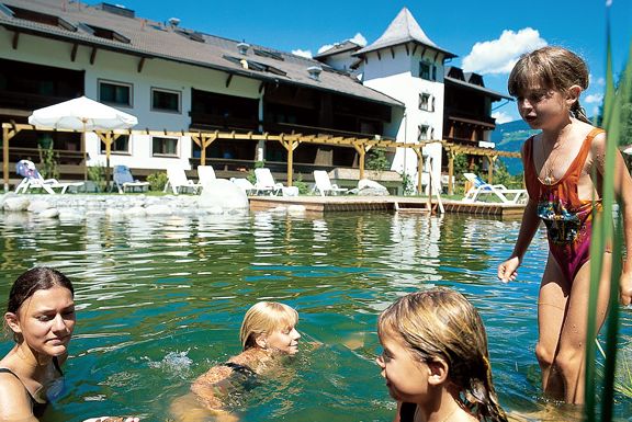 Vier Kinder schwimmen und spielen an einem sonnigen Tag in einem natürlichen Außenpool mit einem Hotelgebäude und Liegestühlen im Hintergrund.
