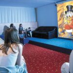 Mehrere Kinder sitzen in einem Raum auf Teppichstufen und dem Fußboden und sehen sich einen Zeichentrickfilm an, in dem Cartoon-Enten auf eine Leinwand projiziert werden.