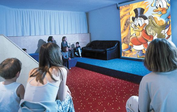 Mehrere Kinder sitzen in einem Raum auf Teppichstufen und dem Fußboden und sehen sich einen Zeichentrickfilm an, in dem Cartoon-Enten auf eine Leinwand projiziert werden.