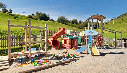 Spielplatz im Freien mit Klettergerüsten, Rutschen, einem Spielhaus und verstreutem bunten Spielzeug auf Sand, eingebettet in eine eingezäunte Rasenfläche mit Hügeln im Hintergrund.