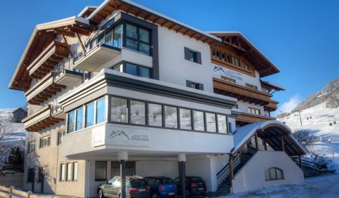 Ein modernes mehrstöckiges Hotel mit Balkonen und hölzernen Akzenten, umgeben von Schnee, mit geparkten Autos davor und einem blauen Himmel darüber.