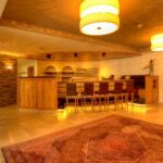 Ein warm beleuchteter Barbereich mit Holzmöbeln, hohen Stühlen, Regalen mit Flaschen und einem großen gemusterten Teppich auf einem gefliesten Boden.