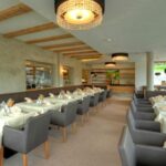 Modernes Restaurantinterieur mit Tischreihen mit weißen Tischtüchern und ordentlich gefalteten Servietten, grauen Stühlen, großen Fenstern und dekorativen Deckenleuchten.