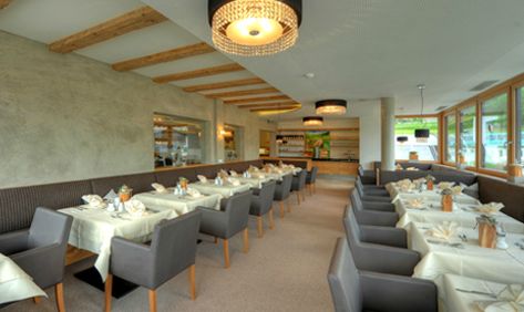 Modernes Restaurantinterieur mit Tischreihen mit weißen Tischtüchern und ordentlich gefalteten Servietten, grauen Stühlen, großen Fenstern und dekorativen Deckenleuchten.