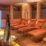 Ein Spa-Entspannungsraum mit mehreren orangefarbenen Liegestühlen, Birkenholzdekor, einer hohen beleuchteten Leuchte und einem Wasserspiel aus Stein in der Mitte.