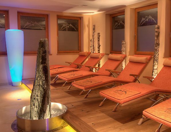 Ein Spa-Entspannungsraum mit mehreren orangefarbenen Liegestühlen, Birkenholzdekor, einer hohen beleuchteten Leuchte und einem Wasserspiel aus Stein in der Mitte.
