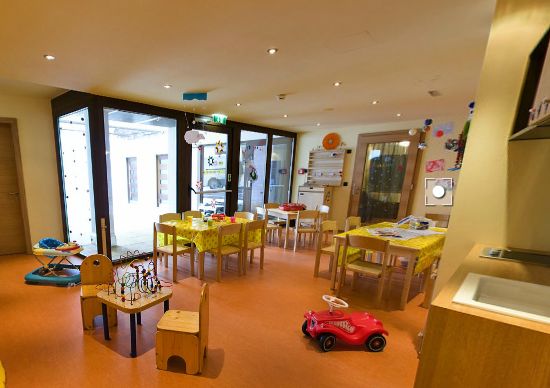 Ein hell erleuchteter Krippenraum mit kleinen Tischen und Stühlen, Kinderspielzeug und Malutensilien, die ordentlich im Raum verteilt sind.