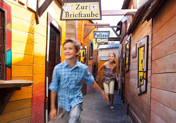 Zwei Kinder rennen lächelnd durch eine bunte Gasse mit Holzhäusern, vorbei an Schildern mit der Aufschrift "Zur Brieftaube" und "Polizei".
