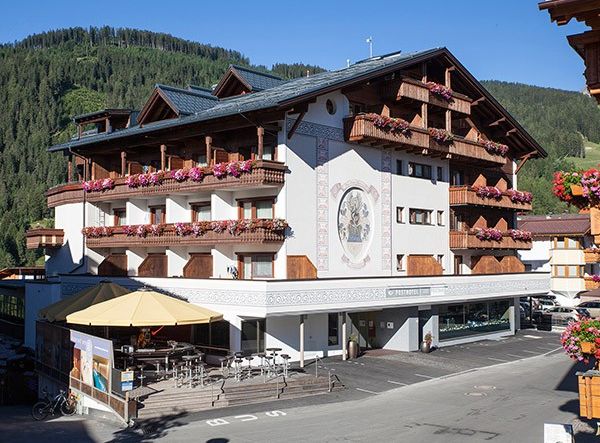 Ein mehrstöckiges Alpenhotel mit Holzbalkonen, die mit rosafarbenen Blumen geschmückt sind, einem Sitzbereich im Freien und einem Wandgemälde an der Fassade vor dem Hintergrund bewaldeter Hügel.