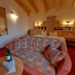 Ein gemütliches Hotelzimmer mit einem gemusterten Sofa, einem Doppelbett, einer Holzdecke, Nachttischen, einem Spiegel und einem kleinen runden Tisch mit einer Flasche, einem Glas und einem Ordner.