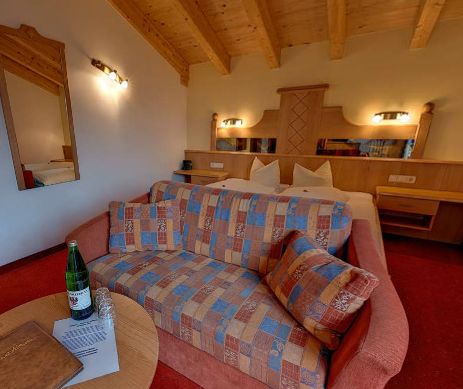 Ein gemütliches Hotelzimmer mit einem gemusterten Sofa, einem Doppelbett, einer Holzdecke, Nachttischen, einem Spiegel und einem kleinen runden Tisch mit einer Flasche, einem Glas und einem Ordner.