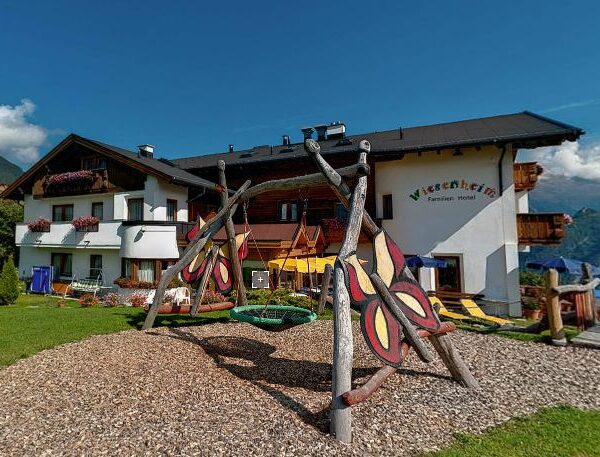 Ein weißes Gästehaus mit Holzbalkonen und Blumenkästen steht hinter einem Spielplatz mit Schaukel auf einer grasbewachsenen Wiese, und im Hintergrund sind die Berge zu sehen.