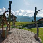 Holzspielplatz Struktur mit Tierkopf Schnitzereien auf einem grasbewachsenen Feld, Berge und Holzgebäude im Hintergrund unter einem teilweise bewölkten Himmel.