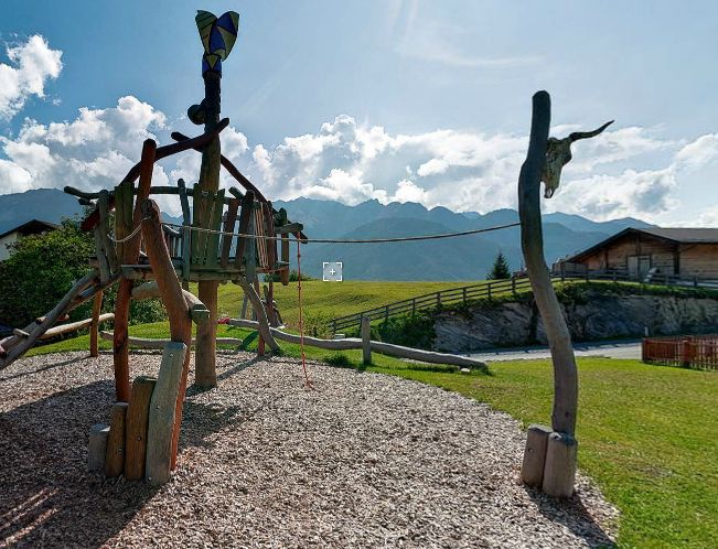 Holzspielplatz Struktur mit Tierkopf Schnitzereien auf einem grasbewachsenen Feld, Berge und Holzgebäude im Hintergrund unter einem teilweise bewölkten Himmel.
