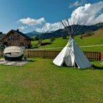 Ein weißes Tipi und ein kleines Boot liegen auf einer Wiese in der Nähe eines Holzhauses, mit Bergen und blauem Himmel im Hintergrund.