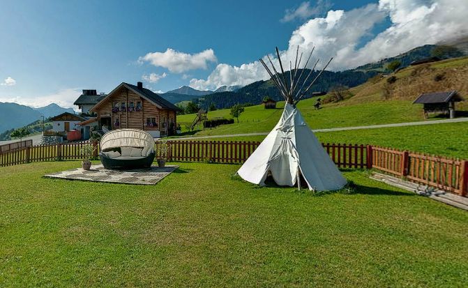 Ein weißes Tipi und ein kleines Boot liegen auf einer Wiese in der Nähe eines Holzhauses, mit Bergen und blauem Himmel im Hintergrund.