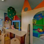 Kinderspielzimmer mit Holztischen und -stühlen, farbenfrohen Spielstrukturen, einem Wandkalender, Regalen mit Spielzeug und Wandbildern mit einer Zeichentrickfigur.