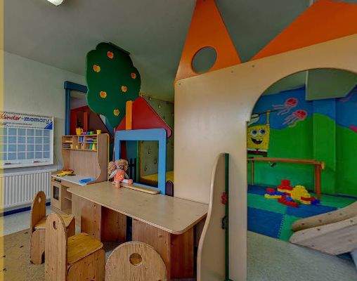Kinderspielzimmer mit Holztischen und -stühlen, farbenfrohen Spielstrukturen, einem Wandkalender, Regalen mit Spielzeug und Wandbildern mit einer Zeichentrickfigur.