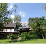 Ein zweistöckiges Haus mit einem dunklen Dach ist von grünen Bäumen und einer Wiese umgeben, mit einem Berg und blauem Himmel im Hintergrund.