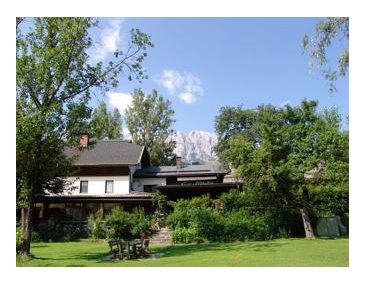 Ein zweistöckiges Haus mit einem dunklen Dach ist von grünen Bäumen und einer Wiese umgeben, mit einem Berg und blauem Himmel im Hintergrund.