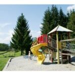 Kinder spielen auf einem Spielplatz mit einer gelben Spiralrutsche und einem Kletternetz, umgeben von Gras, Bäumen und einem blauen Himmel im Hintergrund.