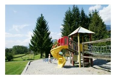 Kinder spielen auf einem Spielplatz mit einer gelben Spiralrutsche und einem Kletternetz, umgeben von Gras, Bäumen und einem blauen Himmel im Hintergrund.