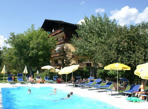 An einem sonnigen Tag faulenzen Menschen auf Stühlen und schwimmen in einem von Bäumen und einem mehrstöckigen Gebäude umgebenen Freibad.