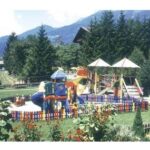 Ein bunter, eingezäunter Spielplatz mit Rutschen und Klettergerüsten befindet sich im Freien, umgeben von Bäumen, Blumen und Bergen im Hintergrund.