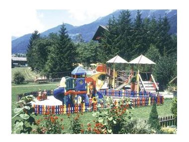 Ein bunter, eingezäunter Spielplatz mit Rutschen und Klettergerüsten befindet sich im Freien, umgeben von Bäumen, Blumen und Bergen im Hintergrund.