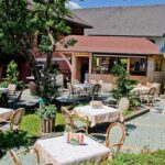 Außencafé mit mehreren Tischen und Stühlen auf einer Steinterrasse, umgeben von Bäumen und Grünflächen, vor einem Holzgebäude.