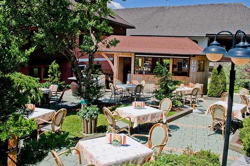 Außencafé mit mehreren Tischen und Stühlen auf einer Steinterrasse, umgeben von Bäumen und Grünflächen, vor einem Holzgebäude.
