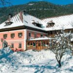 Ein großes rosafarbenes Gebäude mit grünen Fensterläden und einem verschneiten Dach ist umgeben von schneebedeckten Bäumen und Bergen unter einem klaren blauen Himmel.