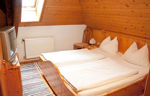 Ein kleines Zimmer im Dachgeschoss mit Holzwänden, zwei zusammengeschobenen Einzelbetten, einem Fernseher, einem Teppich, einem Heizkörper, einem Nachttisch und einer Lampe unter einer schrägen Decke mit einem Dachfenster.