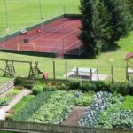 Luftaufnahme eines eingezäunten Gemüsegartens, eines Kinderspielplatzes mit Schaukeln und einer Rutsche und eines eingezäunten Tennisplatzes im Hintergrund.