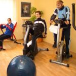 Drei Personen trainieren in einem Fitnessstudio zu Hause an verschiedenen Geräten, darunter ein Liegerad, ein aufrechtes Fahrrad und ein Rudergerät.