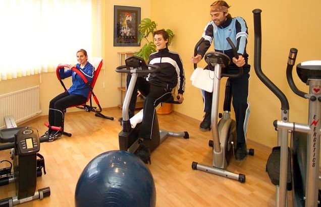 Drei Personen trainieren in einem Fitnessstudio zu Hause an verschiedenen Geräten, darunter ein Liegerad, ein aufrechtes Fahrrad und ein Rudergerät.