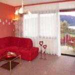 Ein Wohnzimmer mit roten Wänden, einem roten Sektionssofa, einem gläsernen Couchtisch, herzförmiger Dekoration, durchsichtigen Vorhängen und einer offenen Tür, die zu einem Balkon mit Sitzgelegenheiten im Freien und einer malerischen Aussicht führt.