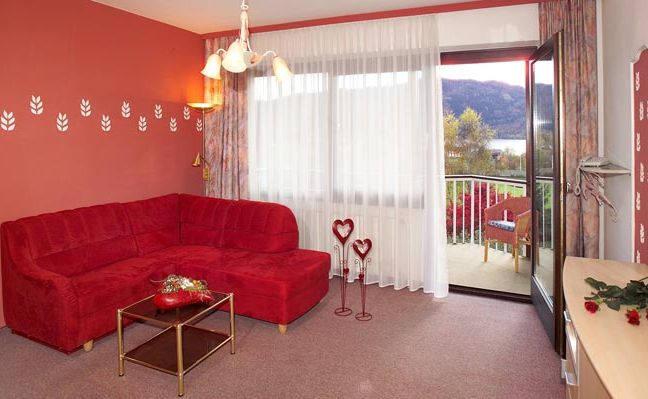 Ein Wohnzimmer mit roten Wänden, einem roten Sektionssofa, einem gläsernen Couchtisch, herzförmiger Dekoration, durchsichtigen Vorhängen und einer offenen Tür, die zu einem Balkon mit Sitzgelegenheiten im Freien und einer malerischen Aussicht führt.