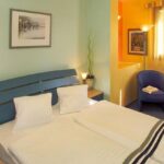 Ein ordentlich gemachtes Doppelbett mit drei Kissen steht in einem hellgrünen Raum, mit zwei Nachttischen, Lampen, einem gerahmten Bild und einer kleinen Sitzecke am Fenster im Hintergrund.