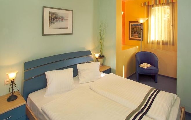 Ein ordentlich gemachtes Doppelbett mit drei Kissen steht in einem hellgrünen Raum, mit zwei Nachttischen, Lampen, einem gerahmten Bild und einer kleinen Sitzecke am Fenster im Hintergrund.