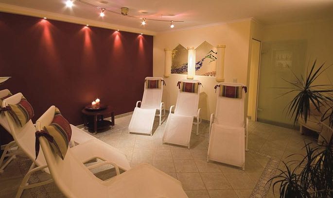 Ein Spa-Entspannungsraum mit fünf weißen Liegestühlen, sanfter Beleuchtung, einem kleinen Tisch mit Kerzen und einer Pflanze in der Ecke. Ein Kunstwerk schmückt die Wand.