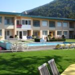 Zweistöckiges Gebäude mit Balkonen, einem Swimmingpool, Liegestühlen und Tischen im Freien auf einer gepflegten Rasenfläche vor der Kulisse grüner Hügel.