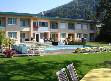 Zweistöckiges Gebäude mit Balkonen, einem Swimmingpool, Liegestühlen und Tischen im Freien auf einer gepflegten Rasenfläche vor der Kulisse grüner Hügel.