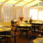 Ein sonnendurchfluteter Essbereich mit mehreren Tischen, die mit gelben und blauen Tischdecken, Tassen, Tellern und Stühlen gedeckt sind, neben großen Fenstern mit durchsichtigen Vorhängen.